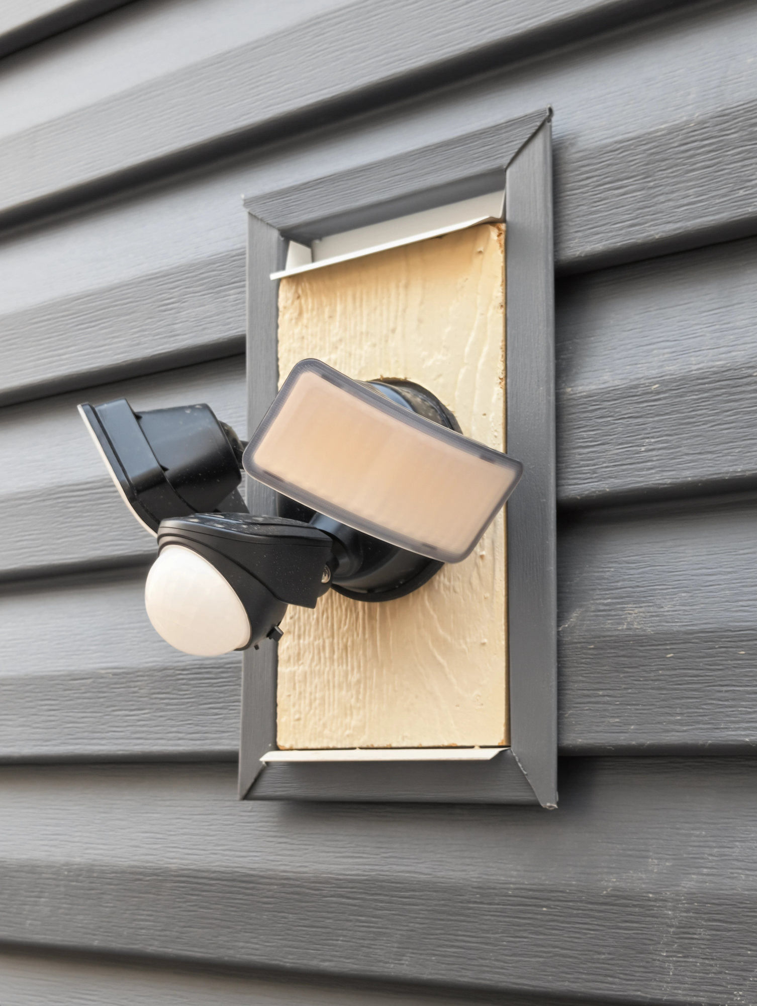 Twin-head motion flood light mounted on a siding block on a dark vinyl garage wall